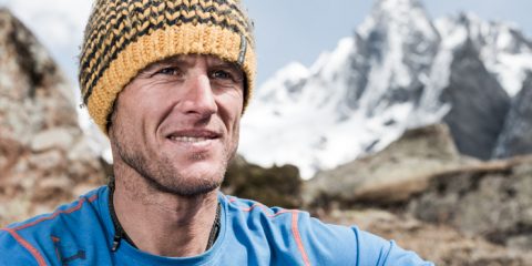 Eine Person mit einer gelb-schwarzen Strickmütze und einer blauen Jacke steht im Freien. Im Hintergrund sind schneebedeckte Berge unter einem bewölkten Himmel zu sehen. Die Person blickt zur Seite und scheint sich in einer Bergregion zu befinden.