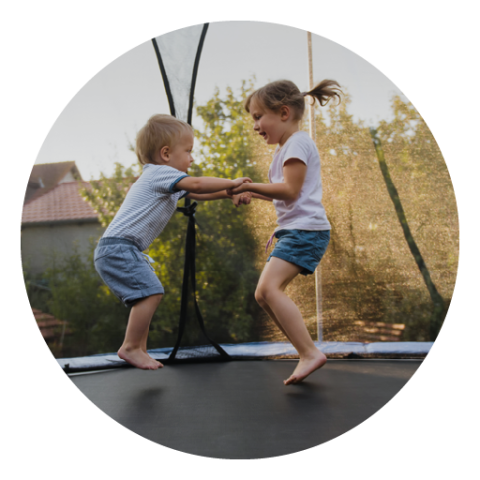 Zwei kleine Kinder spielen fröhlich auf einem Trampolin. Sie halten Händchen und lächeln, umgeben von einem Garten mit Bäumen. Beide tragen Sommerkleidung und im Hintergrund scheint die Sonne.