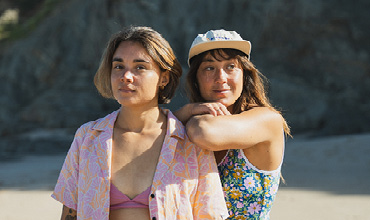 Zwei Frauen stehen dicht beieinander an einem sonnigen Strand. Die eine trägt ein gemustertes Hemd über einem Bikini, die andere ein geblümtes Kleid und eine Mütze, wobei sie ihren Arm auf die Schulter ihrer Freundin legt. Im Hintergrund sind felsige Klippen zu sehen.