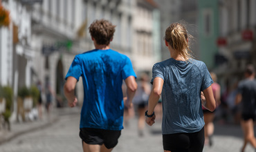 Zwei Personen joggen eine Straße in der Stadt entlang, beide tragen blaue Hemden und schwarze Shorts. Das Foto ist von hinten aufgenommen, und im unscharfen Hintergrund sind andere Personen zu sehen.