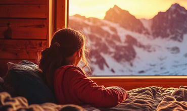 Eine Person mit langen Haaren sitzt auf einem Bett in einem gemütlichen Holzzimmer und blickt aus dem Fenster auf schneebedeckte Berge, die im warmen Licht eines Sonnenauf- oder -untergangs leuchten.