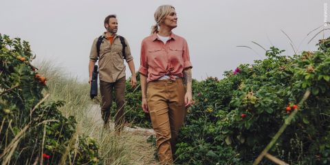 Ein Mann und eine Frau gehen einen Sandweg entlang, der von hohem Gras und grünen Büschen mit Blumen umgeben ist. Beide tragen Straßenkleidung und blicken nach vorne.