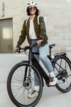 Eine Person mit Helm, Sonnenbrille, grüner Jacke, Jeans und Turnschuhen sitzt lächelnd auf einem schwarzen Fahrrad vor einem hellen Backsteingebäude.