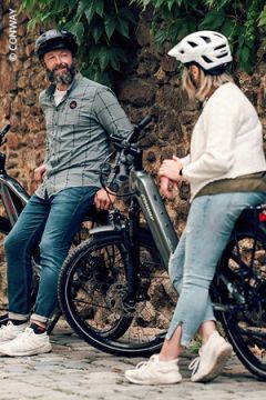 Zwei Menschen mit Helmen und Freizeitkleidung sitzen lächelnd und unterhaltend auf ihren Fahrrädern neben einer begrünten Steinmauer.