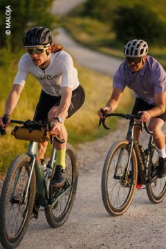 Zwei Radfahrer mit Helmen und Sonnenbrillen fahren auf einem Schotterweg im Grünen. Der vordere Radfahrer trägt ein weißes Hemd und schwarze Shorts, der hintere ein lila Hemd und schwarze Shorts.