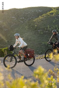 Zwei Radfahrer mit Helmen fahren mit beladenen Tourenrädern eine asphaltierte Straße hinauf durch eine malerische Hügellandschaft, in die das Sonnenlicht einfällt, umgeben von grünen Hügeln und Vegetation.