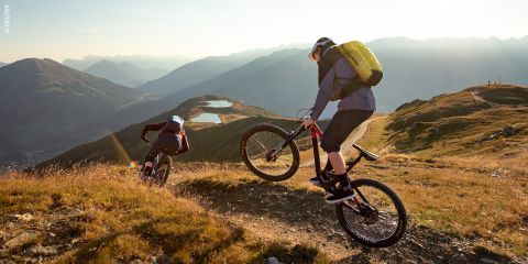 Zwei Mountainbiker fahren auf einem zerklüfteten Weg auf einem sonnenbeschienenen Bergkamm, umgeben von sanften Hügeln, Seen und fernen Berggipfeln unter einem klaren Himmel.
