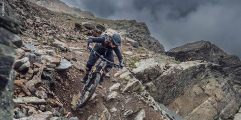 Ein Mountainbiker mit Helm und Ausrüstung fährt einen steilen, felsigen Pfad an einem zerklüfteten Berghang unter einem bewölkten Himmel hinunter.