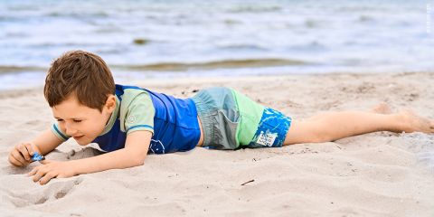Ein kleiner Junge in einem blau-grünen Badeanzug liegt auf dem Bauch im Sand am Strand und spielt mit einem kleinen Spielzeug, während er es genau ansieht. Im Hintergrund ist das Meer zu sehen.