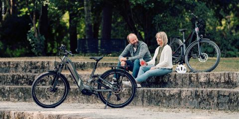 Ein Mann und eine Frau sitzen lächelnd und unterhalten sich auf Steinstufen im Freien. Neben ihnen stehen zwei Elektrofahrräder und ein weißer Helm. Im Hintergrund sind grüne Bäume und Gras zu sehen.