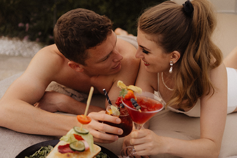 Ein Paar, das im Freien eng beieinander liegt, lächelt und Blickkontakt herstellt, während es bunte Cocktails hält. Im Vordergrund ist Essen zu sehen, und beide wirken entspannt und glücklich.