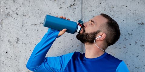 Ein Mann mit einem blauen langärmeligen Hemd und weißen Kopfhörern trinkt aus einer blauen Wasserflasche, während er an einer Betonwand steht.
