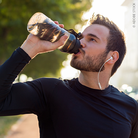 Ein Mann in Sportkleidung trinkt im Freien Wasser aus einer Sportflasche und trägt dabei weiße Kopfhörer. Im Hintergrund scheint das Sonnenlicht, und entlang des Weges sind grüne Bäume zu sehen.