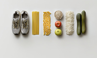 Ein Paar Laufschuhe neben ordentlich angeordneten Lebensmitteln: ungekochte Spaghetti, Penne-Nudeln, Haferflocken, zwei Äpfel, weißer Reis und zwei Zucchini, alle aufgereiht auf einem weißen Hintergrund.