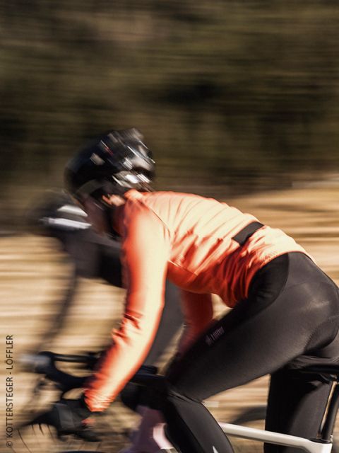 Ein Radfahrer, der eine grüne Löffler-Jacke und einen Helm trägt, schaut mit konzentriertem Blick nach vorn. Das Bild ist leicht unscharf, wodurch die Details des Oberkörpers und der Jacke betont werden.