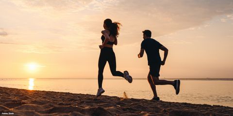 Zwei Personen laufen bei Sonnenaufgang oder Sonnenuntergang an einem Sandstrand entlang, die Silhouette hebt sich vom Himmel ab, das Meer ist im Hintergrund zu sehen. Die heitere Szene fängt ihre Bewegung und die ruhige Atmosphäre des frühen oder späten Tages ein.