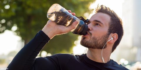 Ein Mann in einem schwarzen langärmeligen Hemd und mit Kopfhörern trinkt an einem sonnigen Tag im Freien Wasser aus einer Sportflasche, mit grünen Bäumen im Hintergrund.