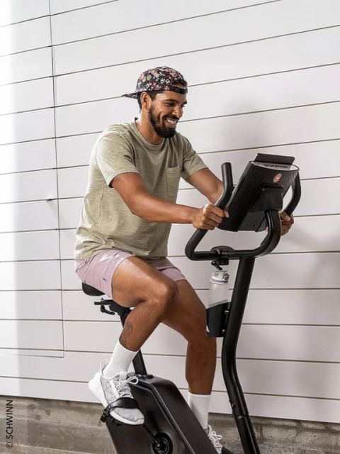 Ein Mann, der eine geblümte Kappe, ein beiges T-Shirt und rosa Shorts trägt, lächelt, während er auf einem stationären Heimtrainer der Marke Schwinn fährt.