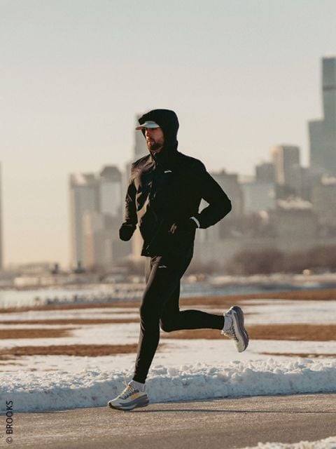 Eine Person, die eine Kapuzenjacke und Laufschuhe trägt, joggt an einem kalten, klaren Tag einen verschneiten Weg entlang, mit der Skyline der Stadt im Hintergrund.