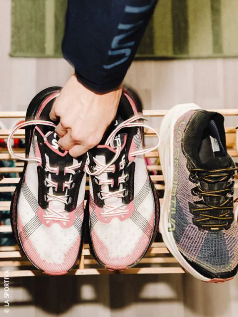 Eine Person hält ein Paar weiß-rosafarbene Laufschuhe an den Schnürsenkeln über einem hölzernen Schuhregal, daneben ein einzelner schwarz-grauer Laufschuh.