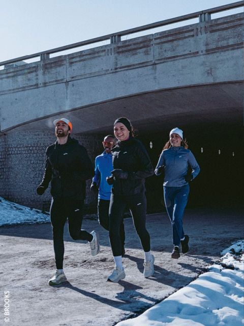 Vier Personen in Winterlaufkleidung joggen gemeinsam auf einem verschneiten Weg unter einer Betonbrücke, lächeln und genießen die Bewegung im Freien.