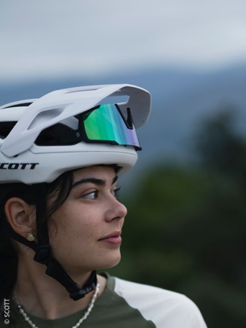 Eine junge Frau mit einem weißen Fahrradhelm und einer reflektierenden Sonnenbrille auf dem Kopf schaut zur Seite. Der Hintergrund ist unscharf mit natürlichem Grün und weichem, bewölktem Himmel.