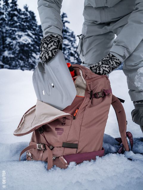 Eine Person in Winterkleidung und gemusterten Handschuhen legt eine Schneeschaufel in einen rosafarbenen Rucksack auf schneebedecktem Boden, mit verschneiten Bäumen im Hintergrund.