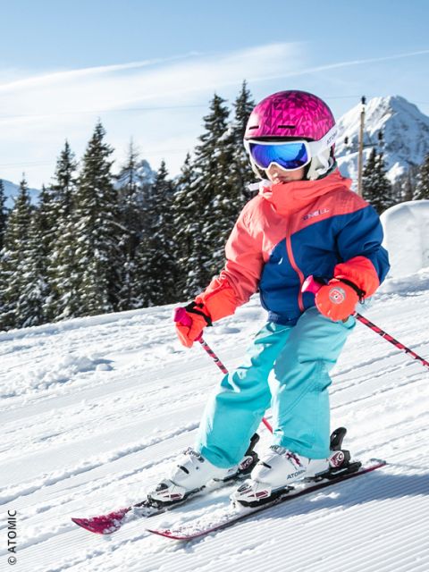 Ein kleines Kind mit einem leuchtend rosa Helm, einer blauen Brille und einer bunten Skiausrüstung fährt auf einer verschneiten Piste bergab, umgeben von Kiefern und Bergen unter einem klaren blauen Himmel.