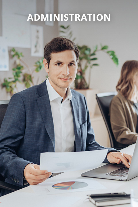 Ein Mann im Anzug sitzt an einem Schreibtisch und hält Dokumente in der Hand, vor ihm befinden sich ein Laptop und Tabellen. Eine Frau arbeitet an einem anderen Schreibtisch im Hintergrund. Oben steht das Wort "ADMINISTRATION".