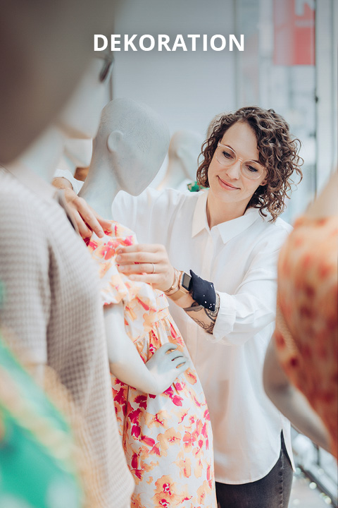 Eine Frau mit lockigem Haar und Brille rückt ein buntes Blumenkleid an einer Schaufensterpuppe in einem hellen Raum zurecht. Das Wort "DEKORATION" erscheint am oberen Rand des Bildes.