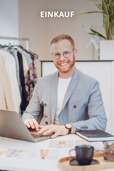 Ein lächelnder Mann mit Brille und einem gestreiften Blazer arbeitet an einem Laptop an einem Schreibtisch mit Modematerialien, einem Notizbuch und einer Kaffeetasse. Im Hintergrund sind Kleiderständer und eine Pflanze zu sehen. Über ihm erscheint das Wort "EINKAUF".