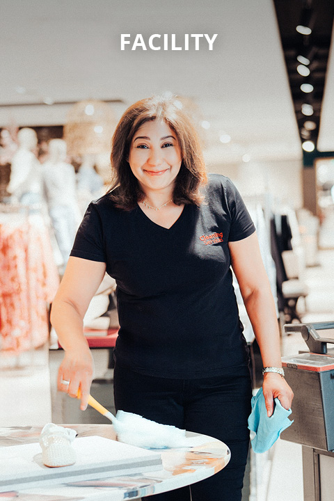 Eine lächelnde Frau in einem schwarzen T-Shirt reinigt einen Tisch mit einem Staubtuch und einem Lappen in einer gut beleuchteten Einrichtung, in der im unscharfen Hintergrund Kleiderständer zu sehen sind. Über ihrem Kopf ist das Wort "FACILITY" zu lesen.