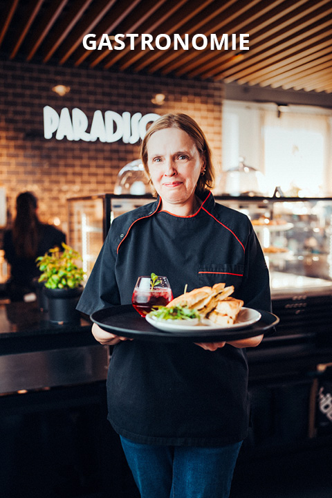 Eine Köchin in schwarzer Uniform hält einen Teller mit Speisen und ein Getränk in einem Restaurant. Über ihr erscheint das Wort "GASTRONOMIE", und im Hintergrund ist ein Schild mit der Aufschrift "PARADISO" zu sehen.