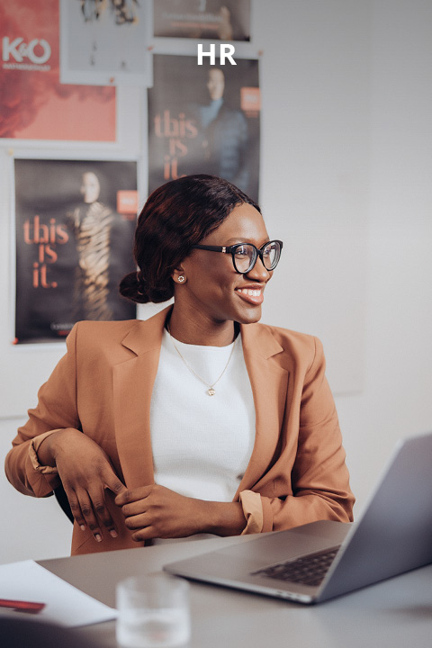 Eine Frau mit Brille und braunem Blazer sitzt an einem Schreibtisch mit einem Laptop, lächelt und blickt zur Seite. An der Wand hinter ihr sind Plakate und das Wort "HR" zu sehen.