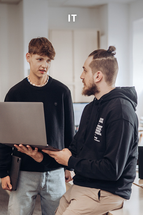 Zwei junge Männer in Freizeitkleidung diskutieren etwas, während sie auf einen Laptop schauen. Einer hält den Laptop und sitzt, während der andere steht. Über ihnen erscheint das Wort "IT", was auf einen informationstechnischen Zusammenhang hinweist.