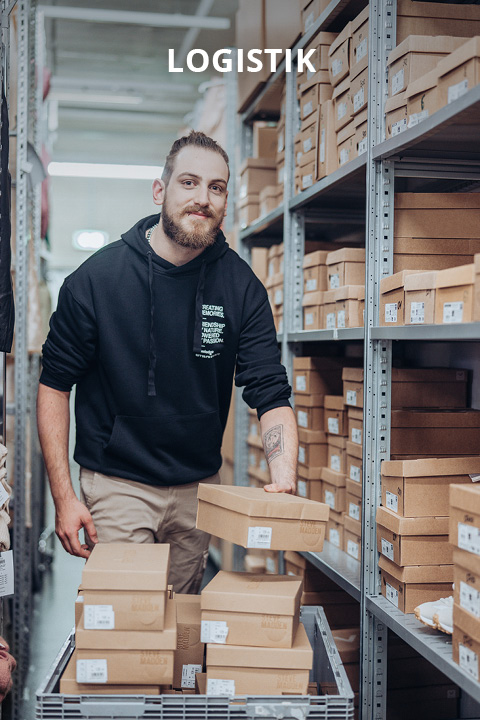 Eine Person mit Bart, die einen schwarzen Kapuzenpulli und eine beigefarbene Hose trägt, verschiebt Kartons auf einem Wagen in einem Lagerraum, der mit Regalen voller ähnlicher Kartons ausgestattet ist. Oben auf dem Bild erscheint das Wort "LOGISTIK".