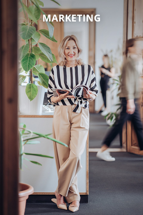 Eine lächelnde Frau in gestreiftem Hemd und beiger Hose steht mit einem aufgeschlagenen Notizbuch in einem modernen Büro mit Pflanzen. Das Wort "MARKETING" erscheint über ihr. Im Hintergrund laufen Menschen vorbei.