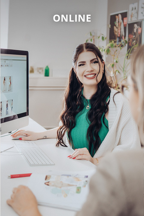 Eine lächelnde Frau mit langen dunklen Haaren sitzt an einem Schreibtisch mit einem Computer und spricht mit einer anderen Person. Auf dem Monitor sind Modebilder zu sehen, und am oberen Rand des Bildes wird das Wort "ONLINE" angezeigt.