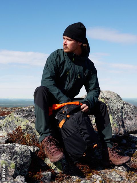 Ein Mann mit dunkler Jacke, schwarzer Mütze und Wanderschuhen sitzt auf felsigem Gelände im Freien und trägt einen schwarz-orangenen Rucksack. Der Himmel ist klar und blau im Hintergrund.