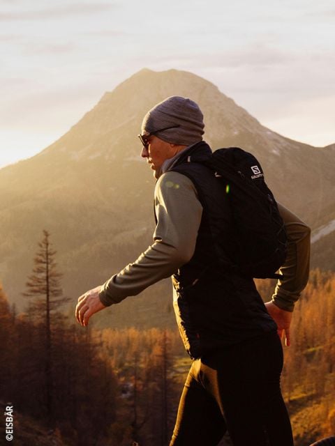 Eine Frau, die eine rosa-graue Jacke und Handschuhe trägt, läuft in einer verschneiten Berglandschaft und trägt zwei Wasserflaschen in den Vordertaschen ihrer Laufweste.