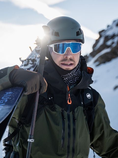 Mann in Winterkleidung und mit Helm hält Skier über die Schulter, trägt eine reflektierende Sonnenbrille und steht in einer verschneiten Berglandschaft.