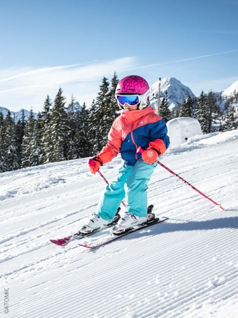 Ein kleines Kind in farbenfroher Skiausrüstung und einem rosa Helm fährt auf Skiern einen verschneiten Hang hinunter, mit Bäumen und Bergen im Hintergrund.