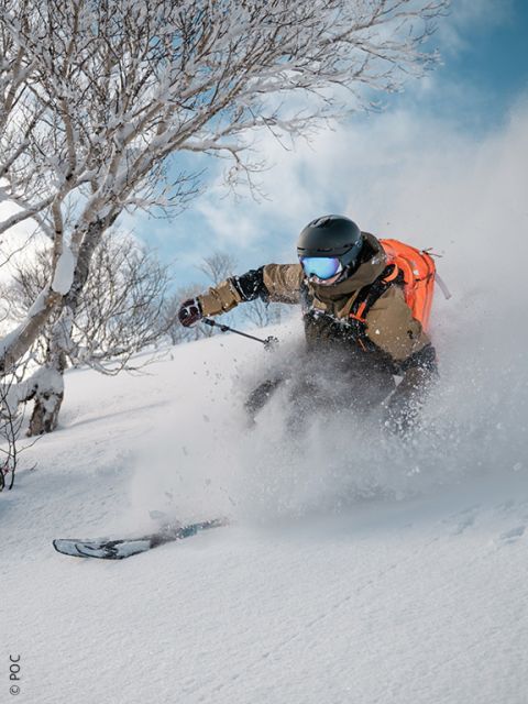 Ein Skifahrer, der einen Helm und einen orangefarbenen Rucksack trägt, fährt durch frischen Pulverschnee und erzeugt eine Gischt, während im Hintergrund schneebedeckte Bäume zu sehen sind.