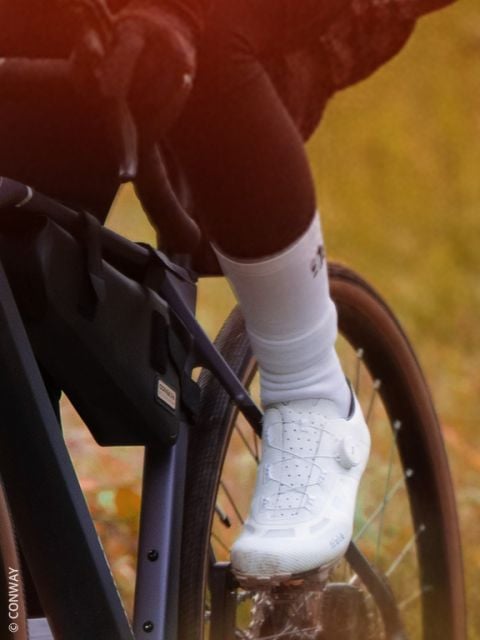 Nahaufnahme des Fußes eines Radfahrers in einem weißen Schuh und einer weißen Socke auf einem Fahrradpedal, mit einem unscharfen Hintergrund aus Gras und gefallenen Blättern.