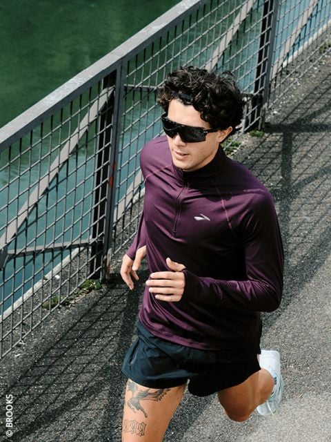 Eine Person, die eine Sonnenbrille und ein lila langärmeliges Oberteil trägt, läuft im Freien neben einem Metallzaun auf einem gepflasterten Weg.