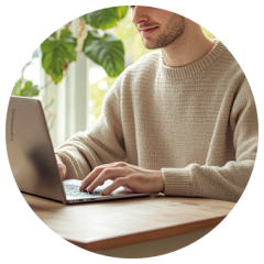 Eine Person in einem beigen Pullover tippt an einem Laptop an einem Holztisch, im Hintergrund sind grüne Pflanzen und Sonnenlicht zu sehen.