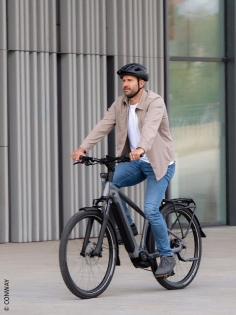 Ein Mann mit Helm und Freizeitkleidung fährt mit einem schwarzen Elektrofahrrad auf einer Straße in der Nähe eines modernen Gebäudes.