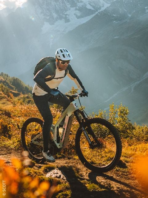 Ein Radfahrer mit Helm und Sonnenbrille fährt auf einem Mountainbike auf einer sonnenbeschienenen Strecke mit Bergen und Grünflächen im Hintergrund.