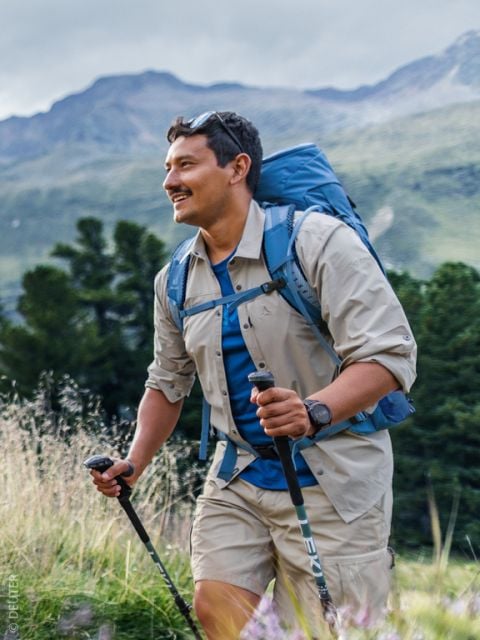 Ein lächelnder Mann mit langen Haaren und einem Bart, der ein blaues Hemd und einen Rucksack trägt, steht im Freien vor einem unscharfen natürlichen Hintergrund.