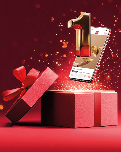 Eine goldene Nummer eins und ein Smartphone tauchen aus einer geöffneten roten Geschenkbox mit einer Schleife vor einem glitzernden roten Hintergrund auf.
